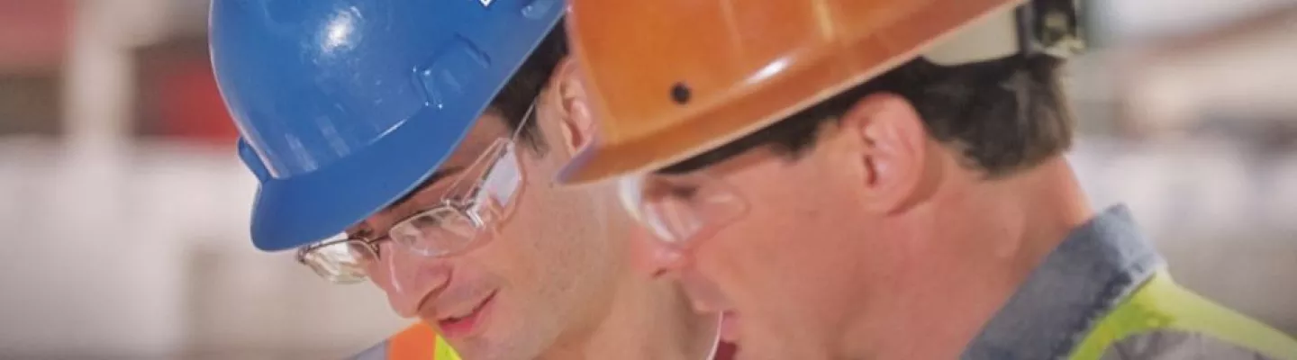 3 men looking at computer screen, one man pointing. All men are wearing hats and safety glasses