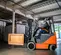 Side view of an orange and grey Toyota warehouse forklift carrying a crate with an operator in an open cabin in a warehouse.