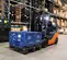 Front angled view of an orange and black forklift lifting blue plastic crates in a warehouse isle driven by an operator.