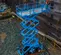 Side top-angled view of a blue scissor lift in use on a construction site, elevated with a worker on the platform.