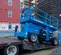 Side view of a blue Genie scissor lift on a trailer ramp at a construction site, with building scaffolding in background.