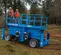 Side view of blue Genie scissor lift with two workers in orange safety gear on the platform on outdoor work zone.