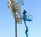 Side angled view of a worker in a blue boom lift lift basket performing maintenance on tall stadium floodlights.