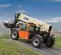 Side view of orange and yellow JLG telehandler with boom retracted and an enclosed cabin on outdoor rough terrain.