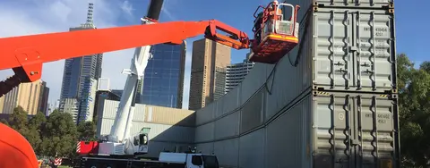 A worker in a cherry picker next to stacked shipping containers, with a large crane and city skyline in the background, indicating temporary worksite facilities.