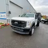 A white Ford truck with a flatbed platform in front of a bright industrial building with a roll-up garage door. The truck is tilted slightly to the left, showing the front and right side.